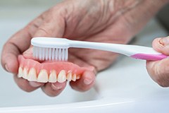 Closeup of someone brushing dentures with a white and pink toothbrush over a sink