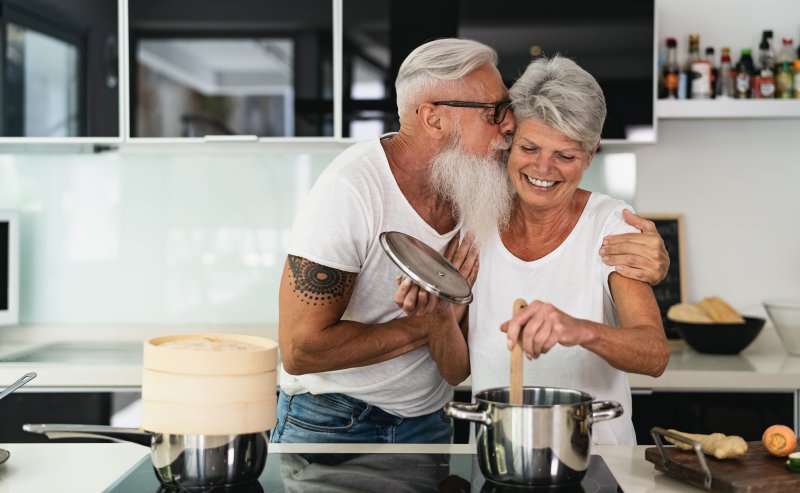 older person with dentures kissing partner on face while cooking